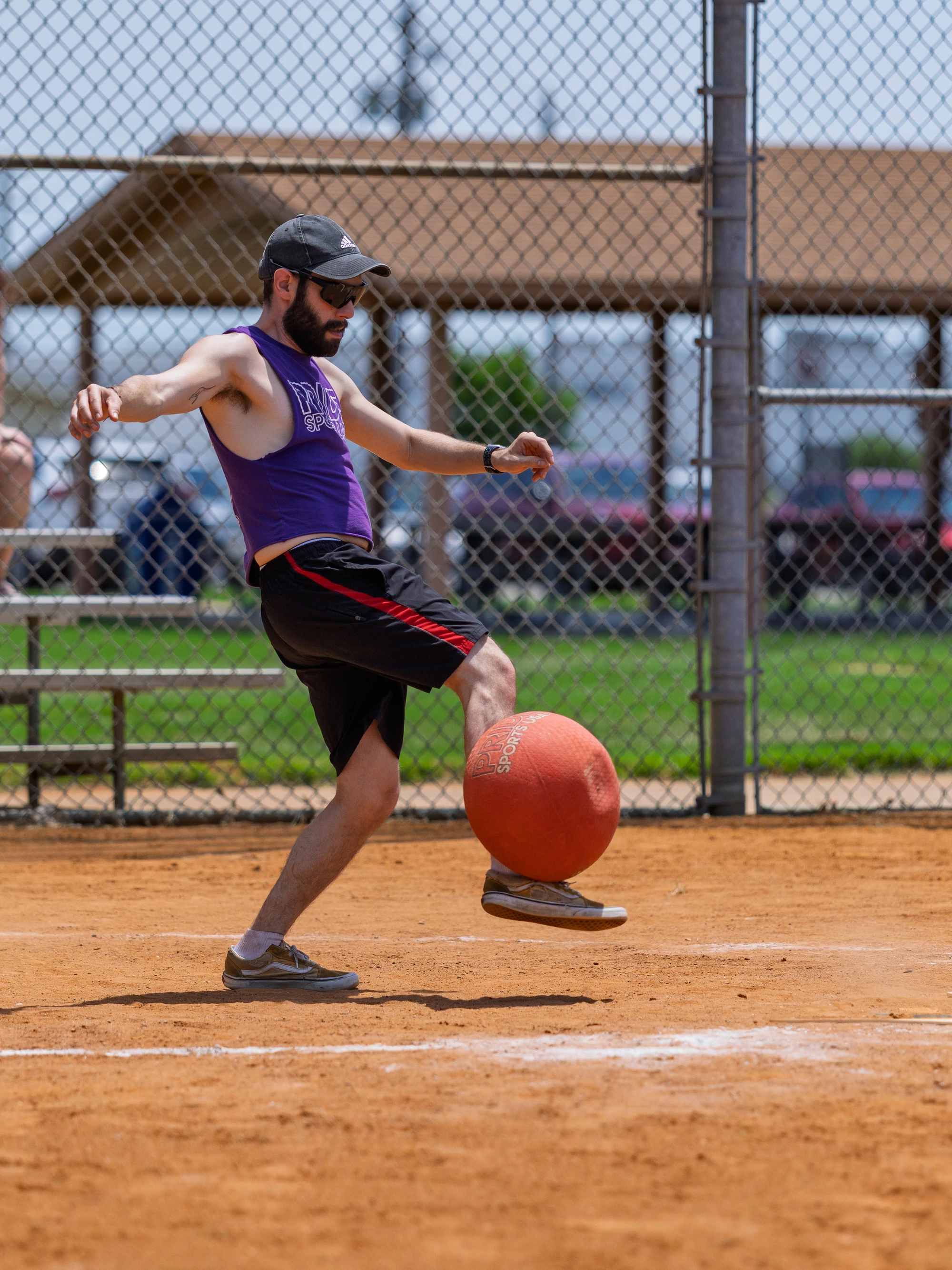A player on a sandy kickball field is wearing a purple tank top, black shorts with red stripes, a cap, and sunglasses, and is in the motion of kicking an orange kickball. There is a chain-link fence and a shelter with benches in the background.