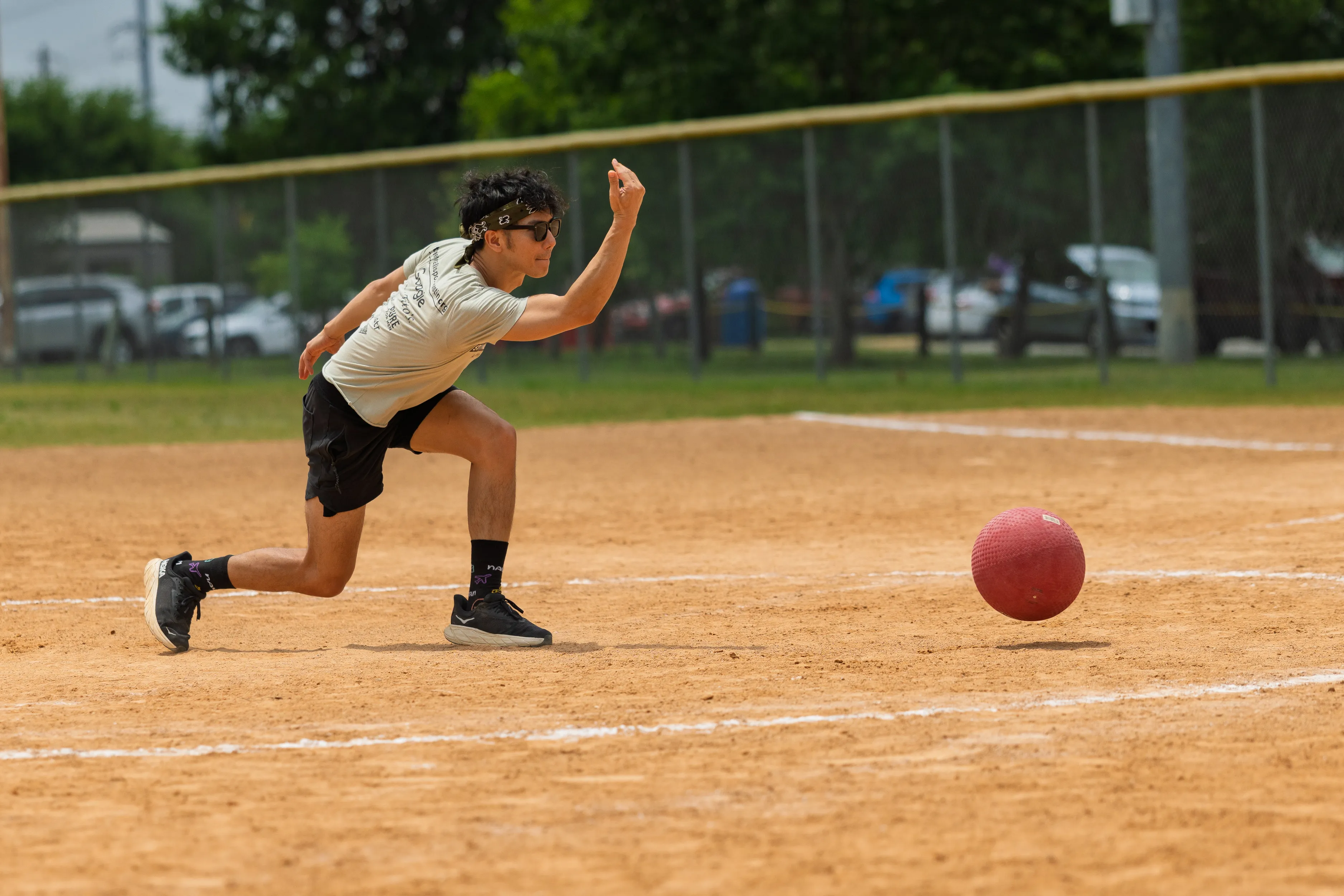 A kickball pitcher in a light T-shirt, black shorts, black socks and sneakers, and a headband lunges low as they release a red kickball down the dusty infield. The play unfolds on a sunny kickball field with chalk baselines, a chain-link fence, and trees in the background.