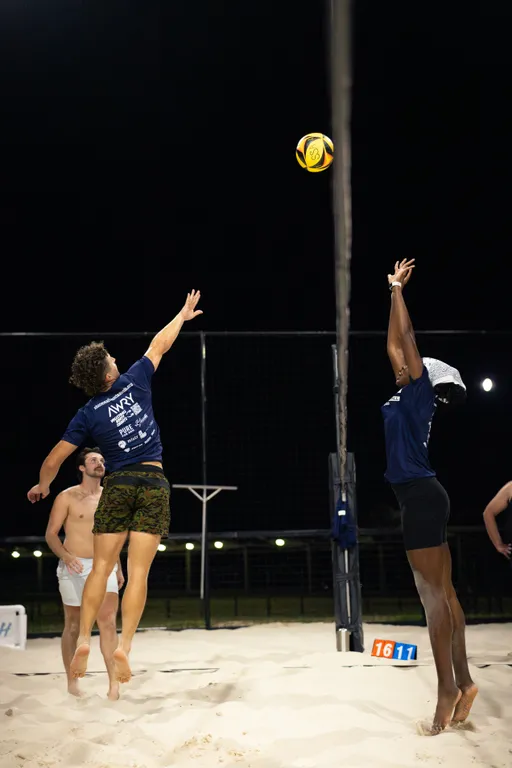 Under stadium lights on a sand volleyball court, two players in navy shirts contest a ball at the net—one jumping in camouflage shorts while the other reaches up to block in black shorts and a white visor as a yellow-and-black volleyball hovers above. Teammates watch from the sand, and a courtside scoreboard shows 16–11.
