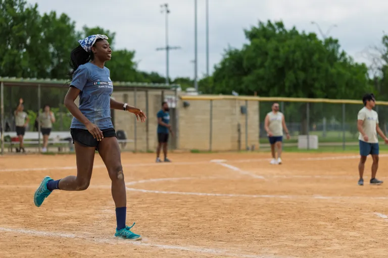 A player wearing a blue shirt, black shorts, blue socks, teal athletic shoes, and a patterned bandana is running on a kickball field, while other players and spectators are visible in the background near the fence and dugout.