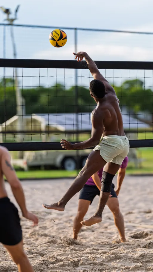 A volleyball player wearing light shorts and a knee brace jumps to spike the ball over the net on a sandy outdoor court, with other athletes and green fields visible in the background.