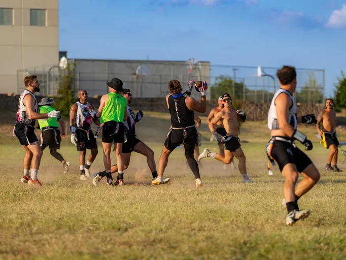 On a sunny grass field, ATX Heat (Black) runs a flag football scrimmage: a receiver reaches up for a pass as defenders converge, kicking up dust with flags trailing from belts. Players wear black practice gear with gloves, headbands, and green pinnies to mark sides, running near a chain-link court and a building in the background.