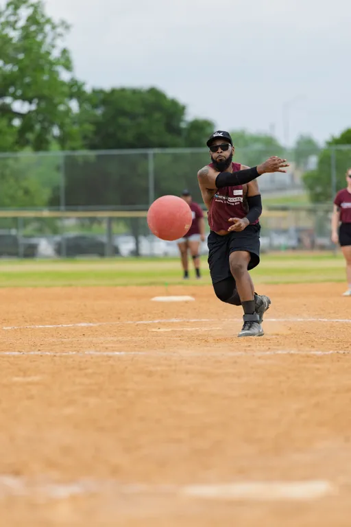 A player on a kickball field is in the process of pitching a red kickball, wearing a maroon team shirt, black shorts, a cap, sunglasses, and knee pads, with teammates visible in the background.