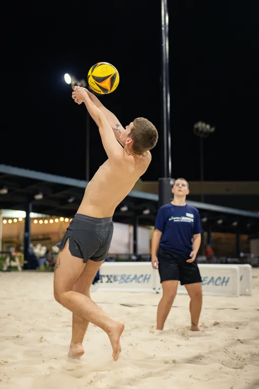 On a lit outdoor sand volleyball court at night, a player in gray shorts leaps with arms extended to bump a yellow-and-black volleyball. A teammate in a navy shirt stands ready behind them near a post, with string lights and court structures in the background.