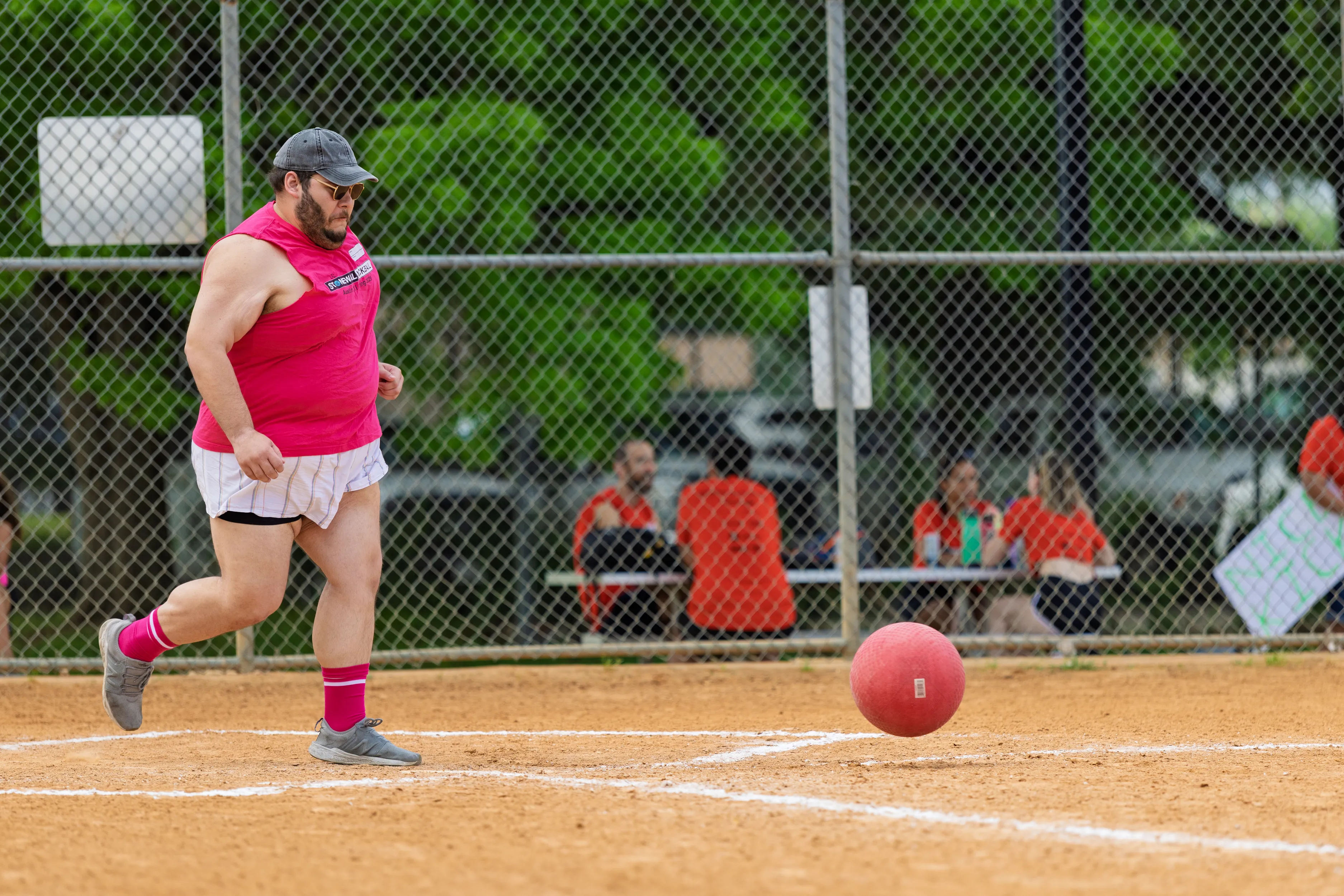 A player wearing a bright pink tank, white skirt over shorts, pink knee-high socks, gray shoes, a cap, and sunglasses runs past home plate as a red kickball rolls along the chalked baseline on a dirt kickball field. Behind a chain-link fence, spectators in orange shirts sit on benches with signs.