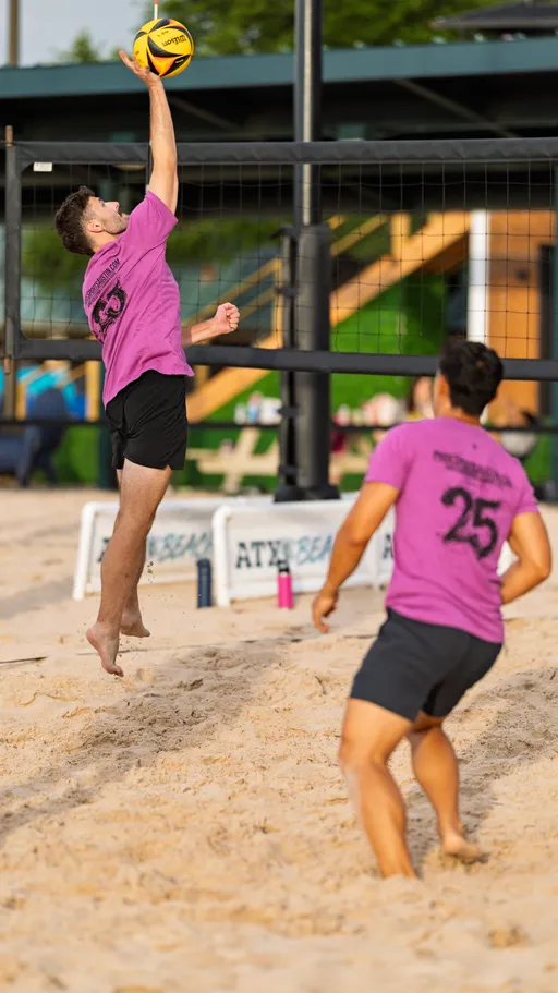 Two volleyball players wearing matching pink shirts and black shorts are competing on a sand court; one player is jumping and reaching high to hit a yellow volleyball over the net, while the other watches and prepares nearby.