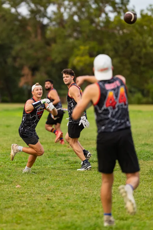 On a grassy park field, ATX Heat (Orange) players in black jerseys with orange trim and “HEAT” text run a flag‑football drill: a quarterback in a white cap throws toward a receiver turning to catch, while teammates in gloves and flag belts track the play against a tree-lined backdrop.