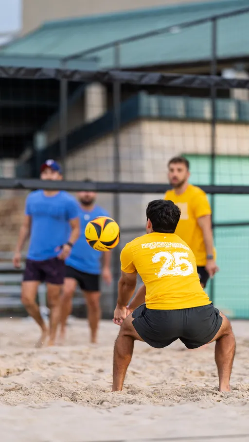 A volleyball player in a yellow shirt and black shorts prepares to receive the ball on a sandy outdoor court, with other players in blue and yellow jerseys visible in the background near the net.