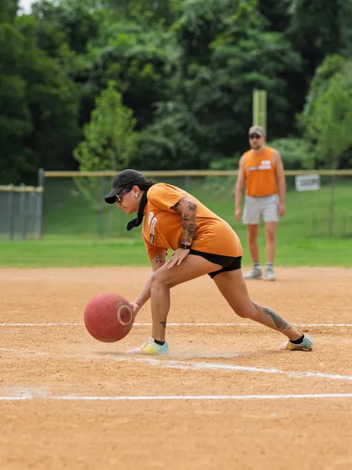 A player wearing an orange shirt, black shorts, and a black cap is crouched low while fielding a red kickball on a dirt kickball field, with another player in orange standing in the background amidst green trees.