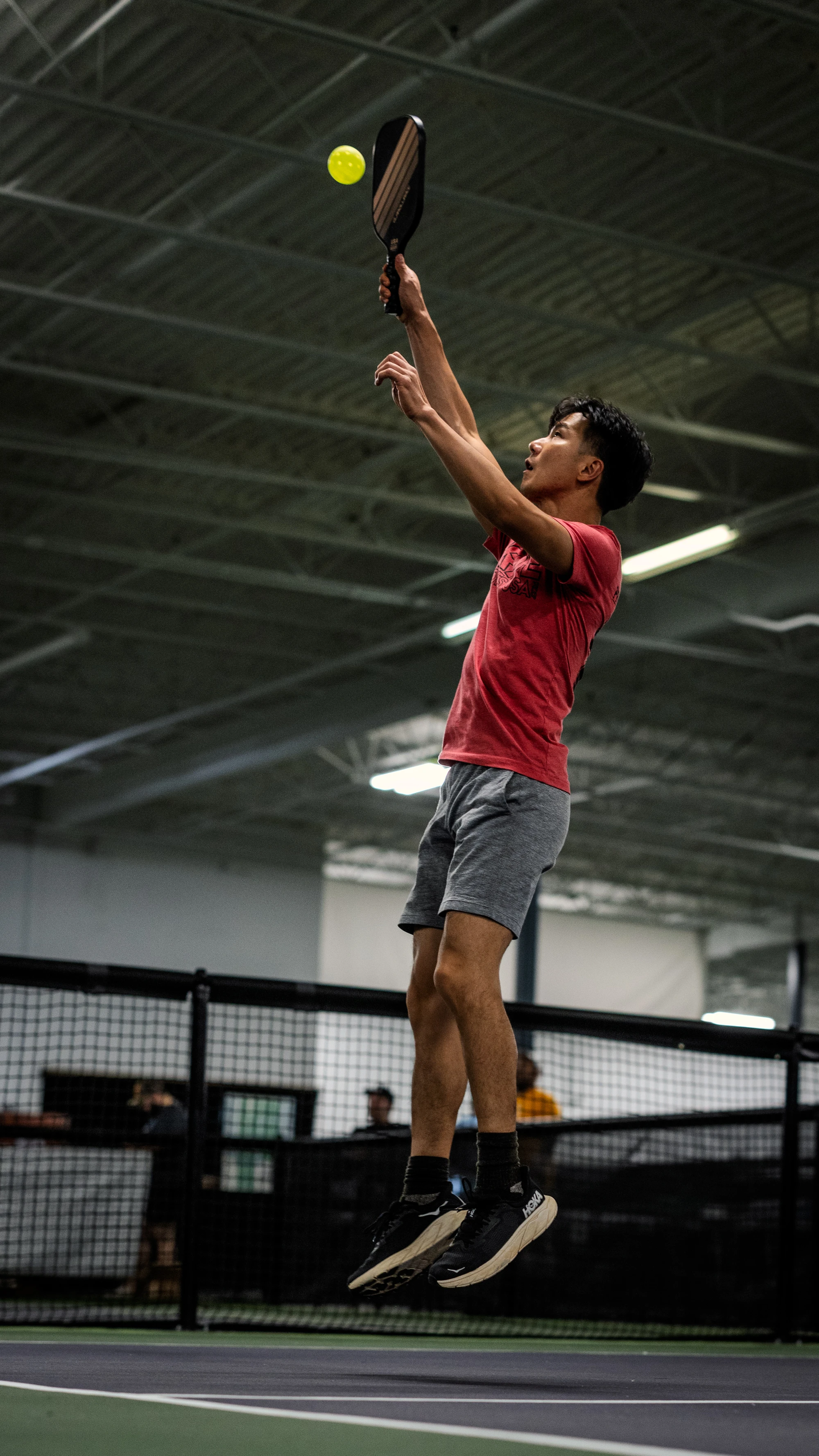 A pickleball player wearing a red shirt and gray shorts is jumping mid-air to hit a yellow pickleball with a paddle on an indoor court.