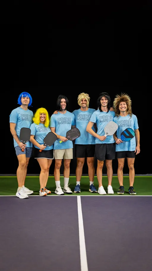 Six players stand side by side on a pickleball court, each holding a paddle and wearing matching blue "Pride Pickleball" shirts and colorful wigs, smiling and posing for a group photo.