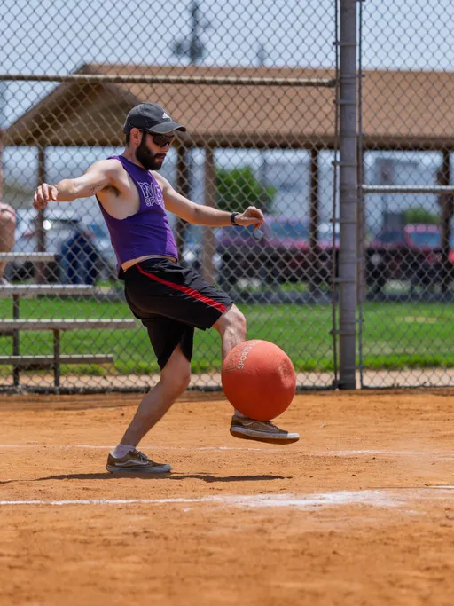 A player on a sandy kickball field is wearing a purple tank top, black shorts with red stripes, a cap, and sunglasses, and is in the motion of kicking an orange kickball. There is a chain-link fence and a shelter with benches in the background.