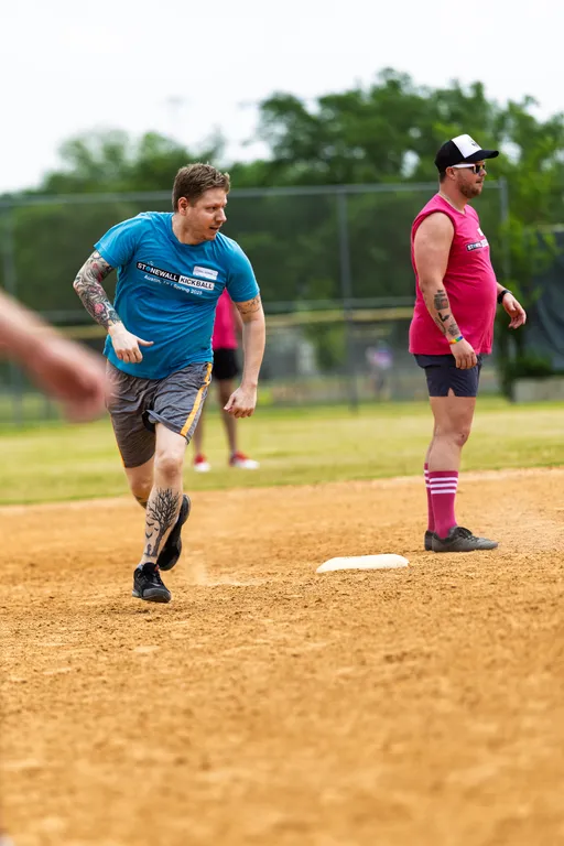A player wearing a blue "Stonewall Kickball" shirt and shorts is running toward a base on a kickball field, while another player in a pink team shirt, matching socks, and a cap stands near the base watching the play.