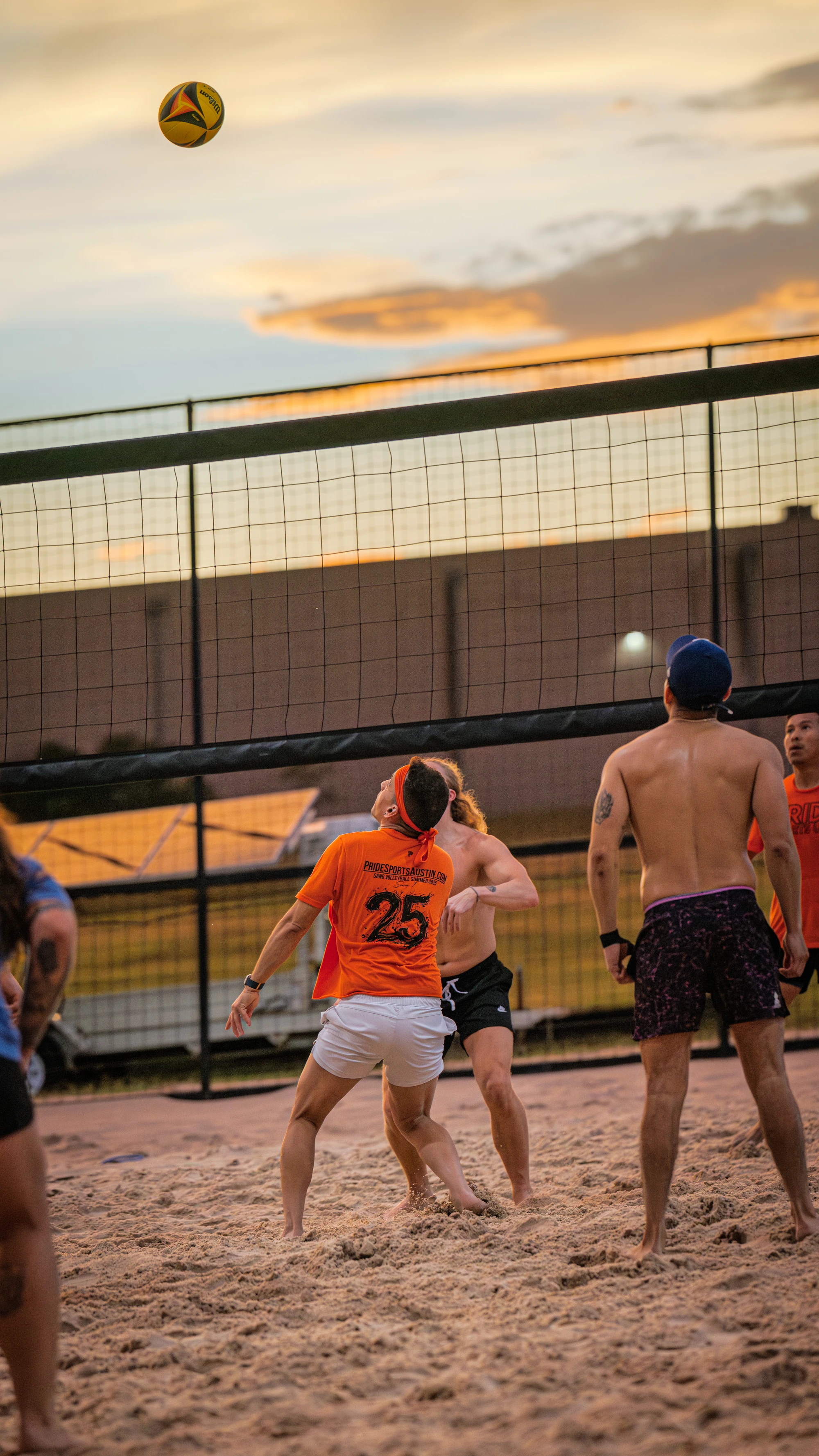 Several volleyball players stand on a sandy court at sunset, watching a volleyball in midair above the net; players wear athletic clothing, including shorts and t-shirts, with some wearing hats or visors.