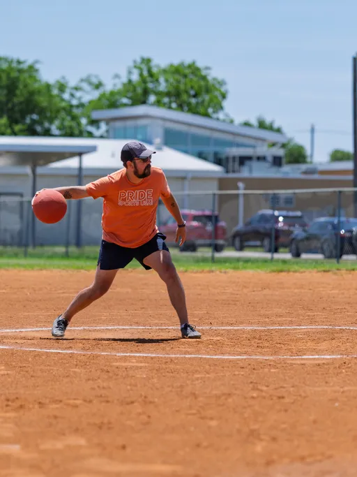 A kickball player wearing an orange "Pride Sports League" shirt, dark shorts, and a cap is winding up to throw a red kickball on a sunny outdoor field. The background shows other field features and parked cars behind a fence.