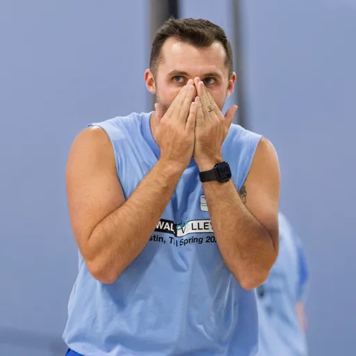 A volleyball player wearing a light blue sleeveless jersey stands on the sand court with hands covering their mouth, appearing focused or surprised during a game.