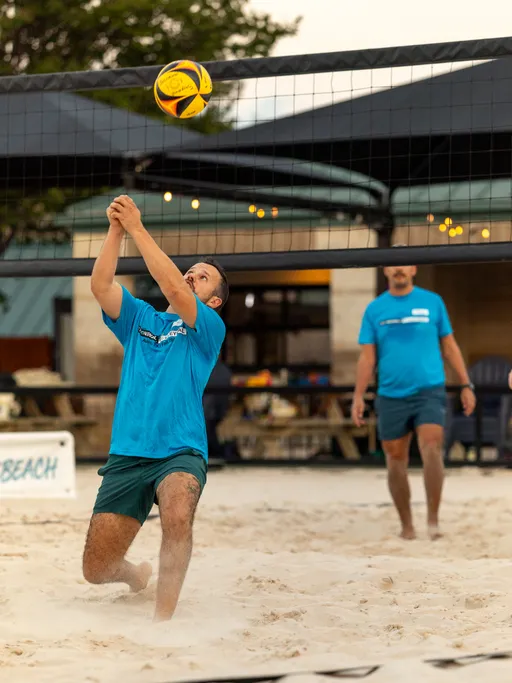 A player in a teal team shirt and dark shorts drops to the sand and raises clasped hands to pass a yellow-and-black volleyball near the net, while a teammate in a matching shirt watches behind. The action is on an outdoor sand volleyball court at dusk, with string lights and a building in the background.