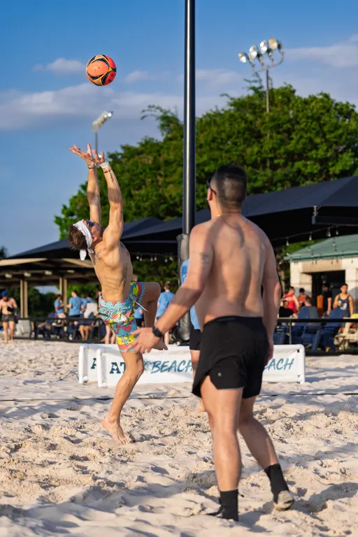 A player jumps to set a volleyball while wearing a headband and colorful shorts, as another competitor stands ready on a sandy outdoor volleyball court with trees and a shaded seating area in the background.