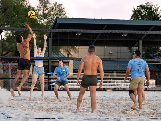 Players on a sand volleyball court are mid-game, with one athlete leaping to spike the ball over the net while another attempts to block; several others in athletic shorts and t-shirts watch and prepare to react, all set against an outdoor recreational facility backdrop.