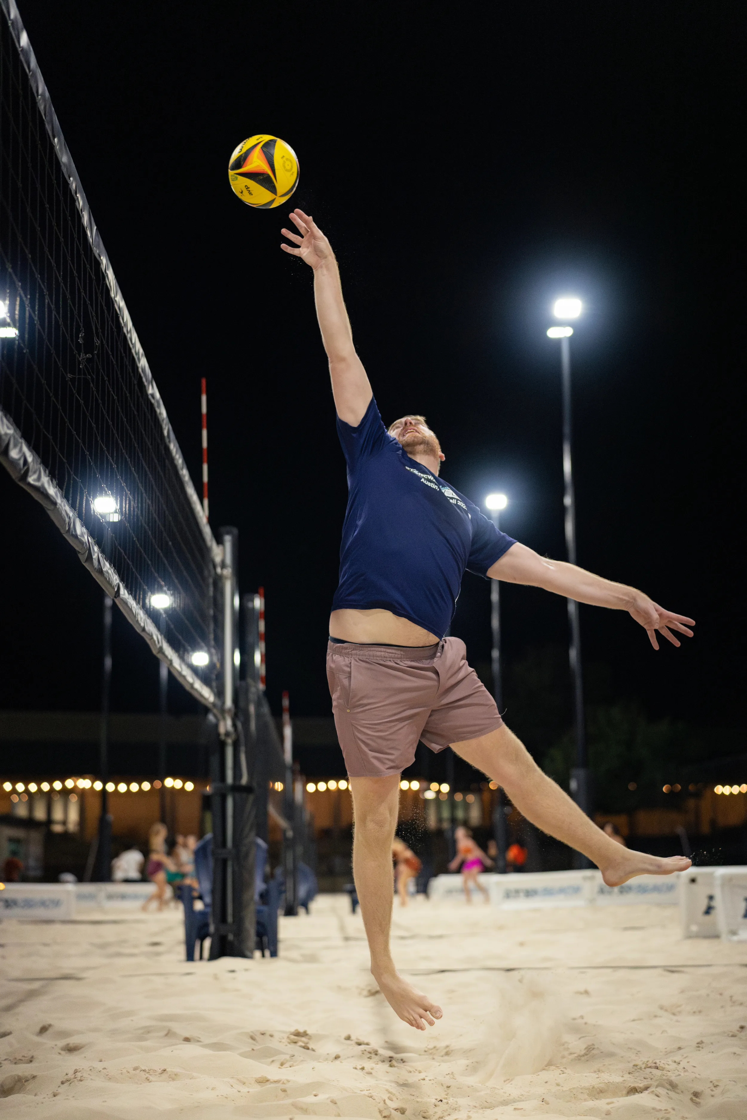 On a lit sand volleyball court at night, a player in a navy shirt and mauve shorts jumps barefoot beside the net, reaching one-handed toward a yellow-and-blue ball for a tip or spike. String lights, tall poles, and other courts with players appear in the background.