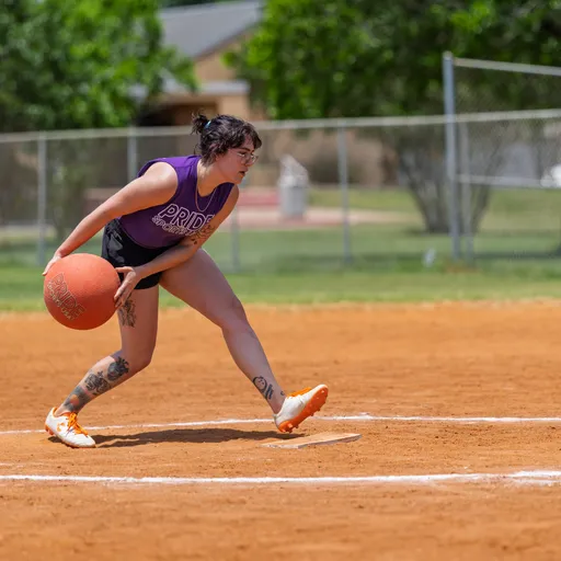 A player wearing a purple "Pride" tank top, shorts, and orange athletic shoes is in motion, preparing to roll a kickball on a dirt field, with a chain-link fence and trees visible in the background.