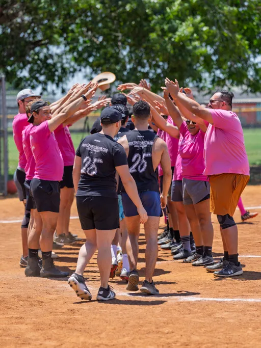 Players wearing bright pink and dark uniforms form two lines on a kickball field, creating an arch with their raised arms while others walk underneath, celebrating together in a festive outdoor setting.