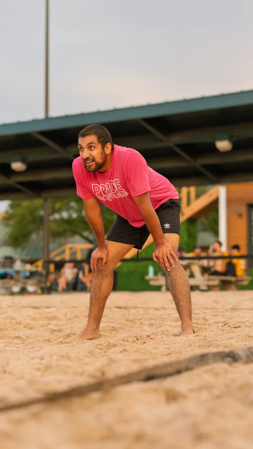 A volleyball player wearing a bright pink "Pride Sand Volleyball" shirt and black shorts is crouched and ready in a defensive stance on a sandy court, with an outdoor pavilion and spectators visible in the background.