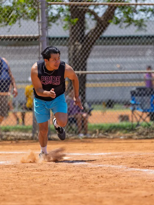 A player wearing a sleeveless "Pride Kickball USA" shirt and light blue shorts is sprinting across a dusty kickball field, with a chain-link fence and trees visible in the background.