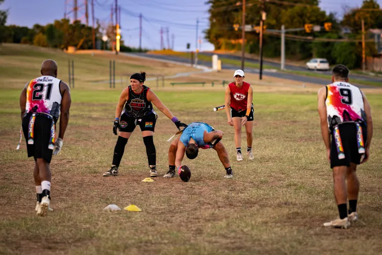 On a grassy field at dusk, ATX Heat (Black) athletes run a flag‑football snap drill: a center in a light‑blue top bends to hike the ball between cones while teammates in black HEAT jerseys set to rush or receive, and players in multicolor‑numbered practice jerseys space out along the line. Flag belts, cleats, gloves, and compression gear are visible, with an open park, road, and trees in the background.