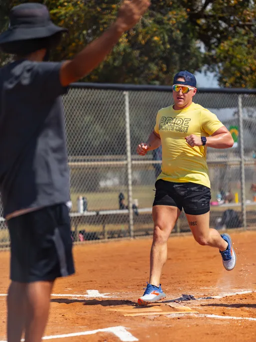 On a sunny kickball field, a player in a yellow “PRIDE SPORTS” shirt, black shorts, blue shoes, sunglasses, and a backward cap sprints across the home plate line, kicking up dust. A teammate or coach in a dark outfit and wide-brim hat raises both arms near the baseline, with a chain-link fence and trees in the background.