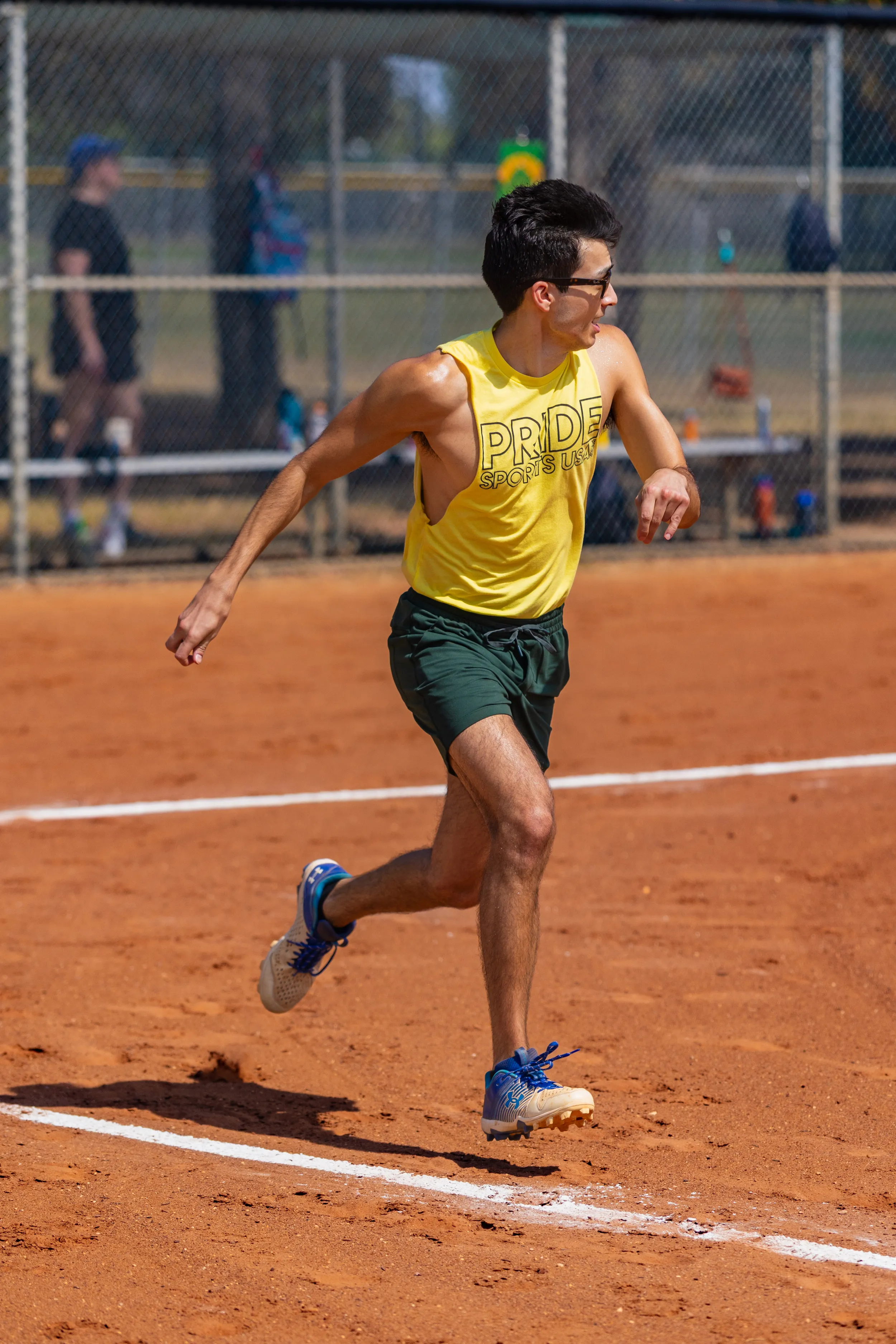 A kickball player in a yellow “PRIDE SPORTS” tank, dark green shorts, blue sneakers, and sunglasses runs along a white-chalk baseline on a red-dirt kickball field, looking toward the infield. A chain-link fence and other players are blurred in the background.