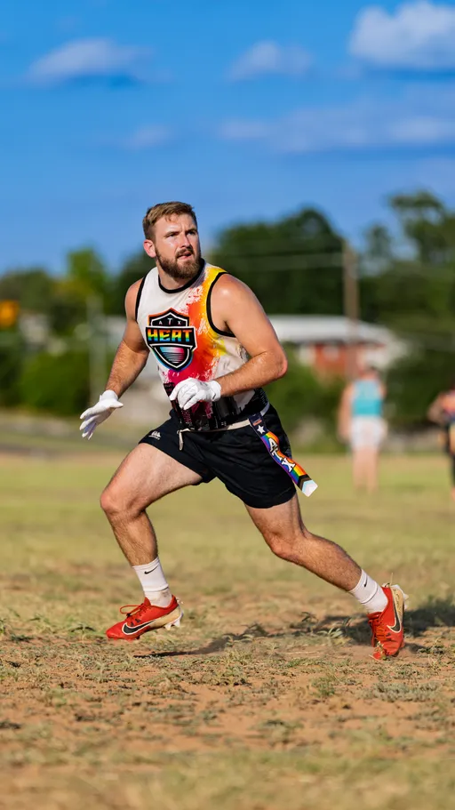 An ATX Heat (Black) flag football player runs on a sunny practice field, looking upfield, wearing a black jersey with a colorful HEAT logo, black shorts with a rainbow stripe, white gloves, red cleats, and a belt with pull flags. Blurred teammates and trees sit in the background during Gay Bowl 2025 practice.