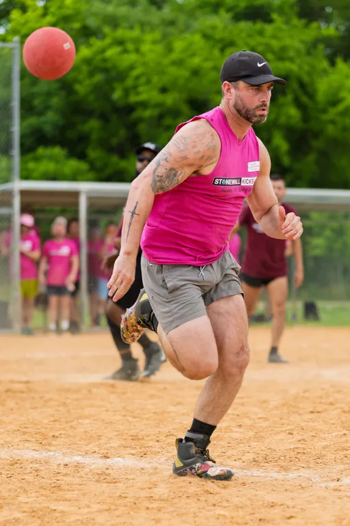 A kickball player in a pink sleeveless Stonewall Kickball shirt and gray shorts is in motion on a dirt field, appearing to have just kicked a red kickball, with other players and spectators visible in the background.
