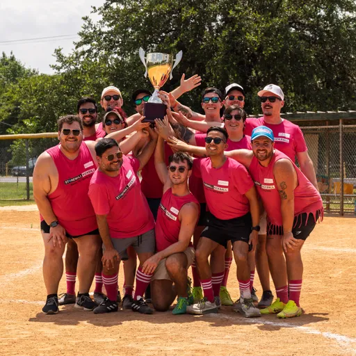 A team of kickball players wearing matching bright pink shirts, athletic shorts, and striped knee-high socks pose together on a dirt kickball field, holding up a large trophy and smiling in celebration.