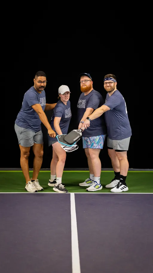 Four pickleball players stand together on an indoor court, each wearing matching athletic shirts and shorts, holding their paddles in the center with their hands stacked in a team gesture. The group appears ready and unified, with the paddle court lines visible in the foreground.