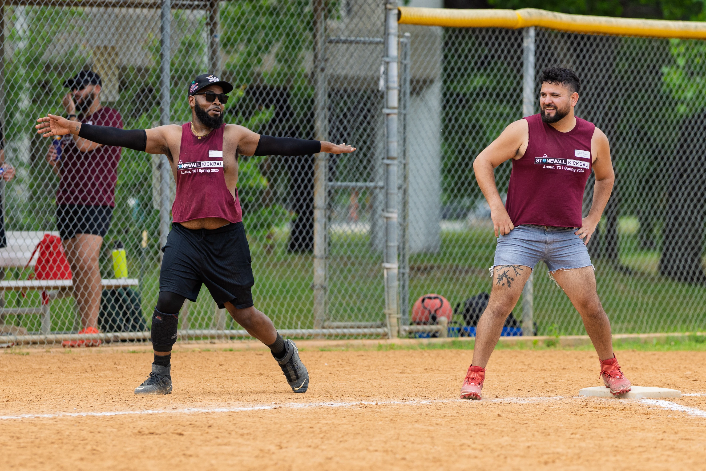 Two kickball players wearing matching maroon Stonewall Kickball shirts are on a dirt field near a base; one player is mid-stride with arms extended, while the other stands with hands on hips and a tattoo on one leg. A chain-link fence, additional players, and sports equipment can be seen in the background.
