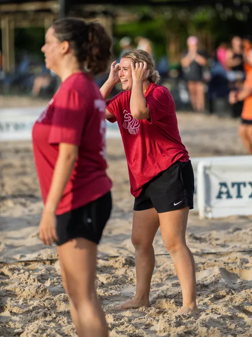 Two volleyball players wearing matching red shirts and black shorts stand on a sandy volleyball court, with one player appearing to call out or signal while holding hands near their face. The background shows more players and a partially blurred environment, indicating active gameplay and a lively match setting.