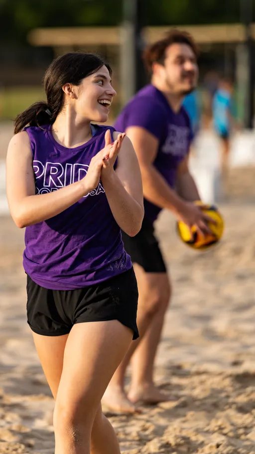 Two volleyball players in matching purple "Pride" shirts and black shorts are on a sandy court; one athlete is holding a volleyball while another claps and smiles, suggesting excitement during a match.