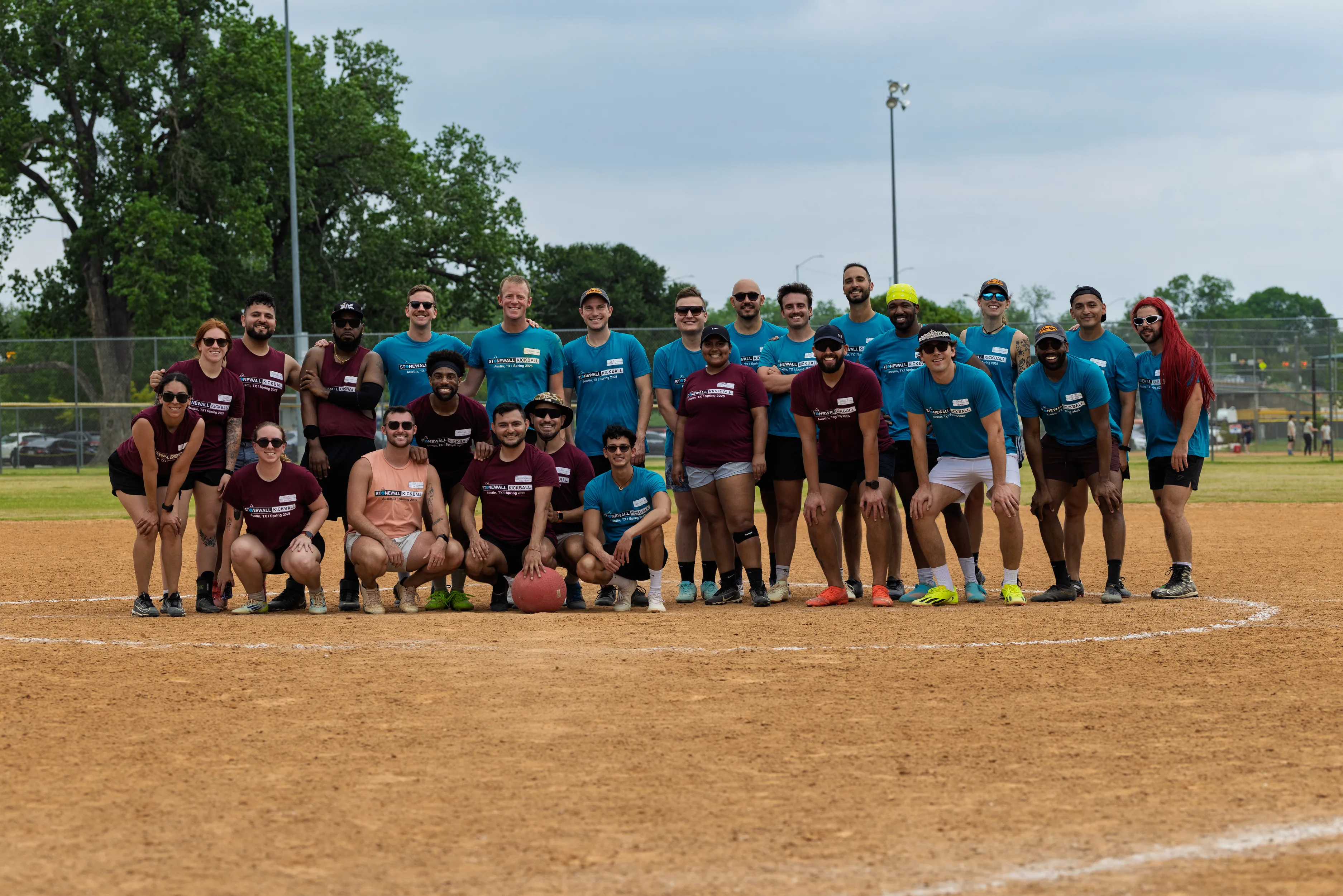 Two groups of kickball players in matching jerseys—maroon and teal—pose together at home plate on a dirt kickball field; some kneel while others stand, one holding a red kickball, with chain-link fencing, trees, and field lights in the background.