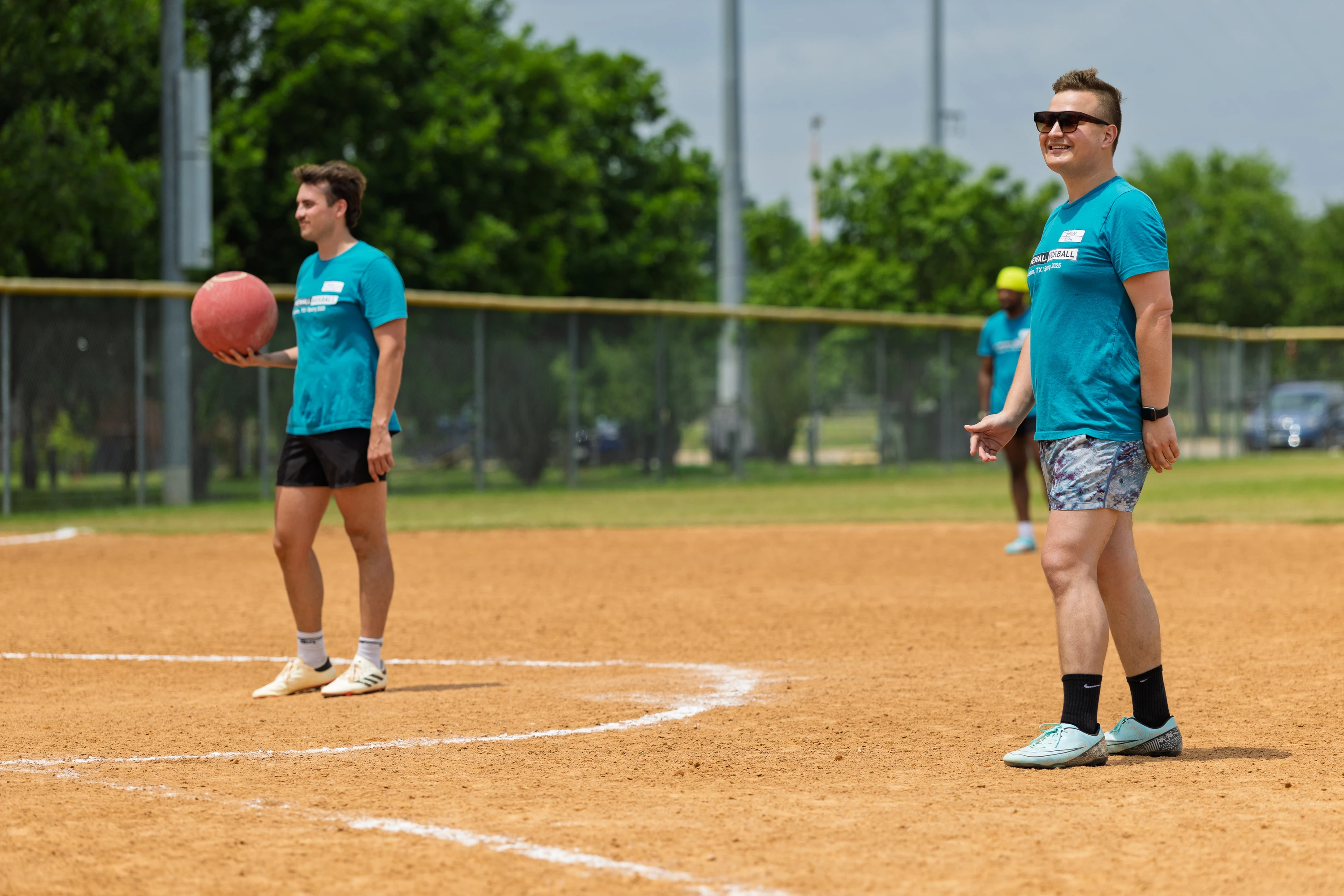 Two kickball players wearing matching teal shirts and shorts stand on a dirt field; one player is holding a red kickball while the other smiles and faces forward, with trees and a fence visible in the background.