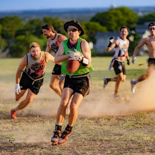 ATX Heat (Black) players scrimmage on a dry, grassy field during Gay Bowl 2025 practice; a ball carrier in a green mesh pinnie, hat, and sunglasses sprints with the football while defenders in ATX Heat jerseys, shorts, and flag belts chase through a cloud of dust against a tree-lined hillside.