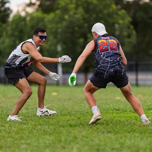 During ATX Heat’s Gay Bowl practice, two players run a flag‑football drill on a grassy field: a defender in sunglasses and gray gloves crouches with arms wide, closing on a player in a black‑and‑gray jersey with orange number 22, backward cap, and bright green glove who plants to cut away; yellow cones mark the training area with a fence and trees in the background.