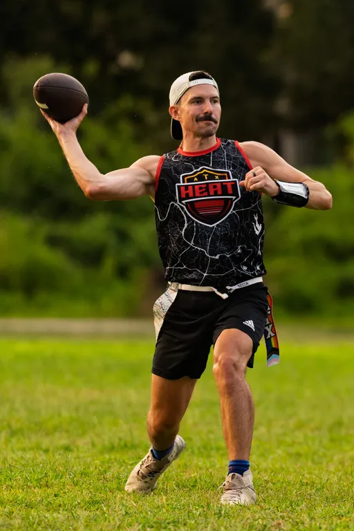On a grassy field, an ATX Heat (Orange) flag football player winds up to throw a pass, wearing a black sleeveless jersey with the Heat logo, black shorts, cleats, a backward white cap, a wrist play band, and a rainbow-striped flag belt; trees blur in the background during practice for Gay Bowl 2025.