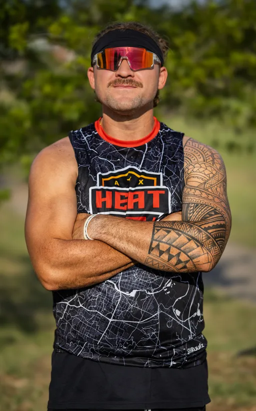 An ATX Heat (Black) athlete stands on a grassy practice field preparing for Gay Bowl 2025, arms crossed, wearing a black sleeveless jersey with a city-map pattern and bold ATX HEAT logo, mirrored sport sunglasses, and a headband. A geometric arm tattoo and wristband are visible with blurred trees in the background.