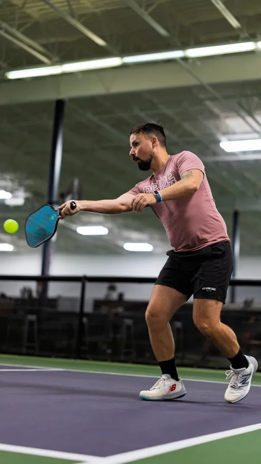 A pickleball player in athletic clothing and sneakers is preparing to hit a ball with a paddle on an indoor court, wearing a "PRIDE" shirt during a match.
