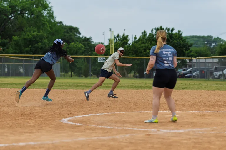 Three players are on a kickball field; one player is running toward a red kickball in midair, another player is extending an arm as if throwing or catching, and a third stands nearby watching. All wear athletic clothing and sneakers, and the scene takes place on a dirt infield surrounded by grass and trees.