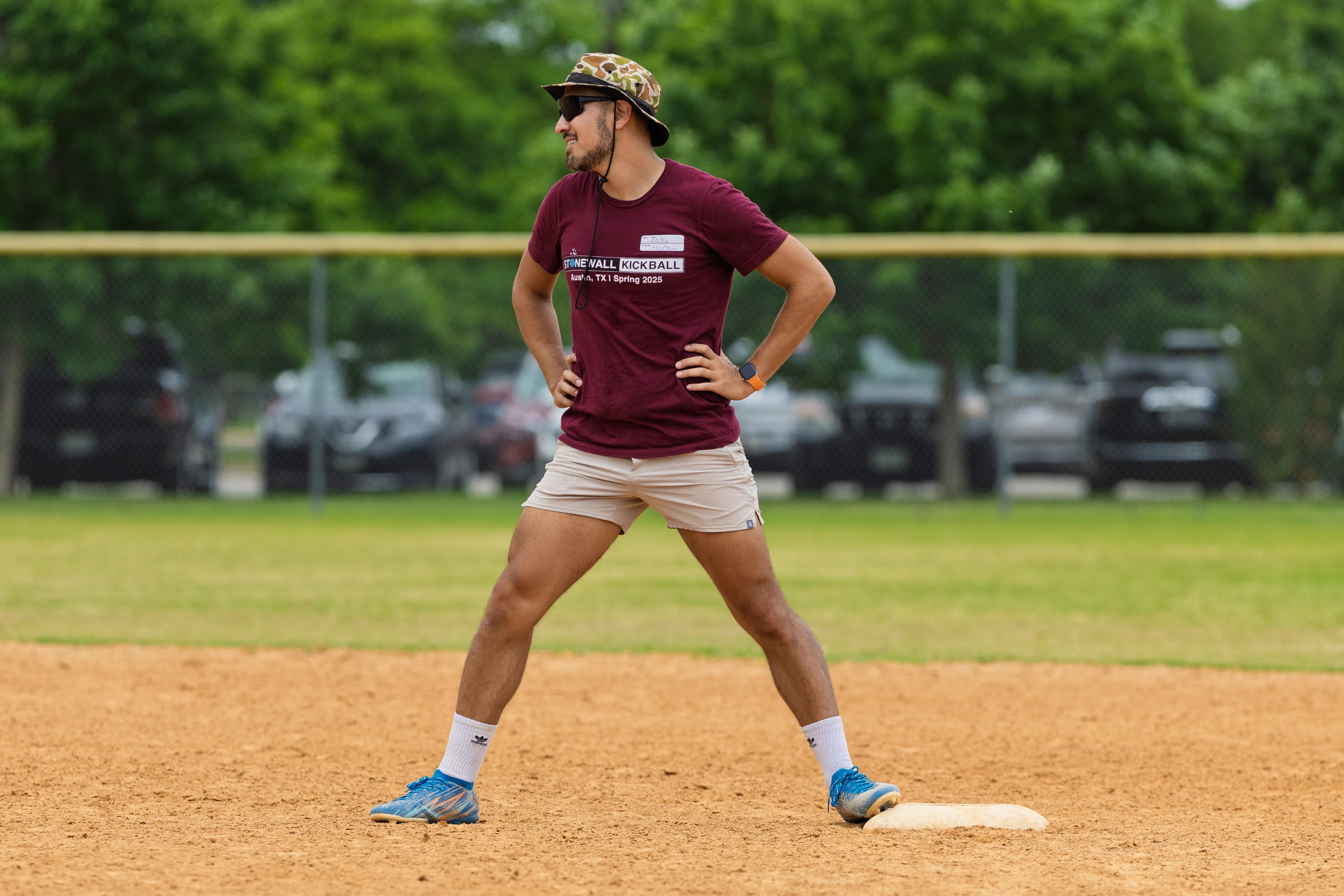 A kickball player stands on a base with hands on hips, wearing a maroon "Stonewall Kickball Spring 2025" shirt, khaki shorts, blue athletic shoes, a patterned bucket hat, and sunglasses on a sandy kickball field with trees and cars in the background.