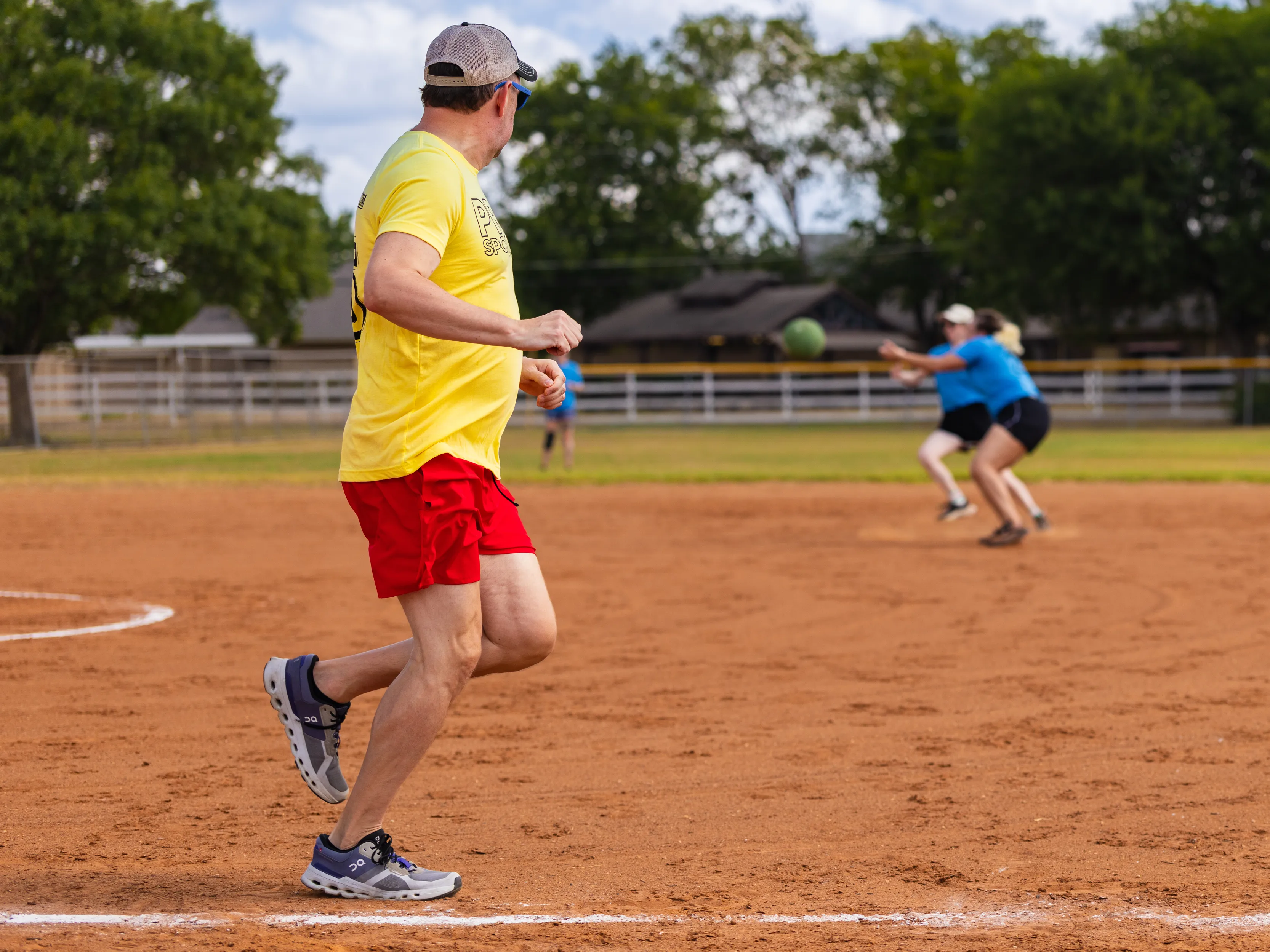 On a sunny kickball field, a player in a yellow shirt and red shorts stands near the baseline watching a green kickball fly toward two fielders in blue shirts who are moving in to catch it. The scene is set on a red-dirt infield with white chalk lines, a fence, and trees in the background.