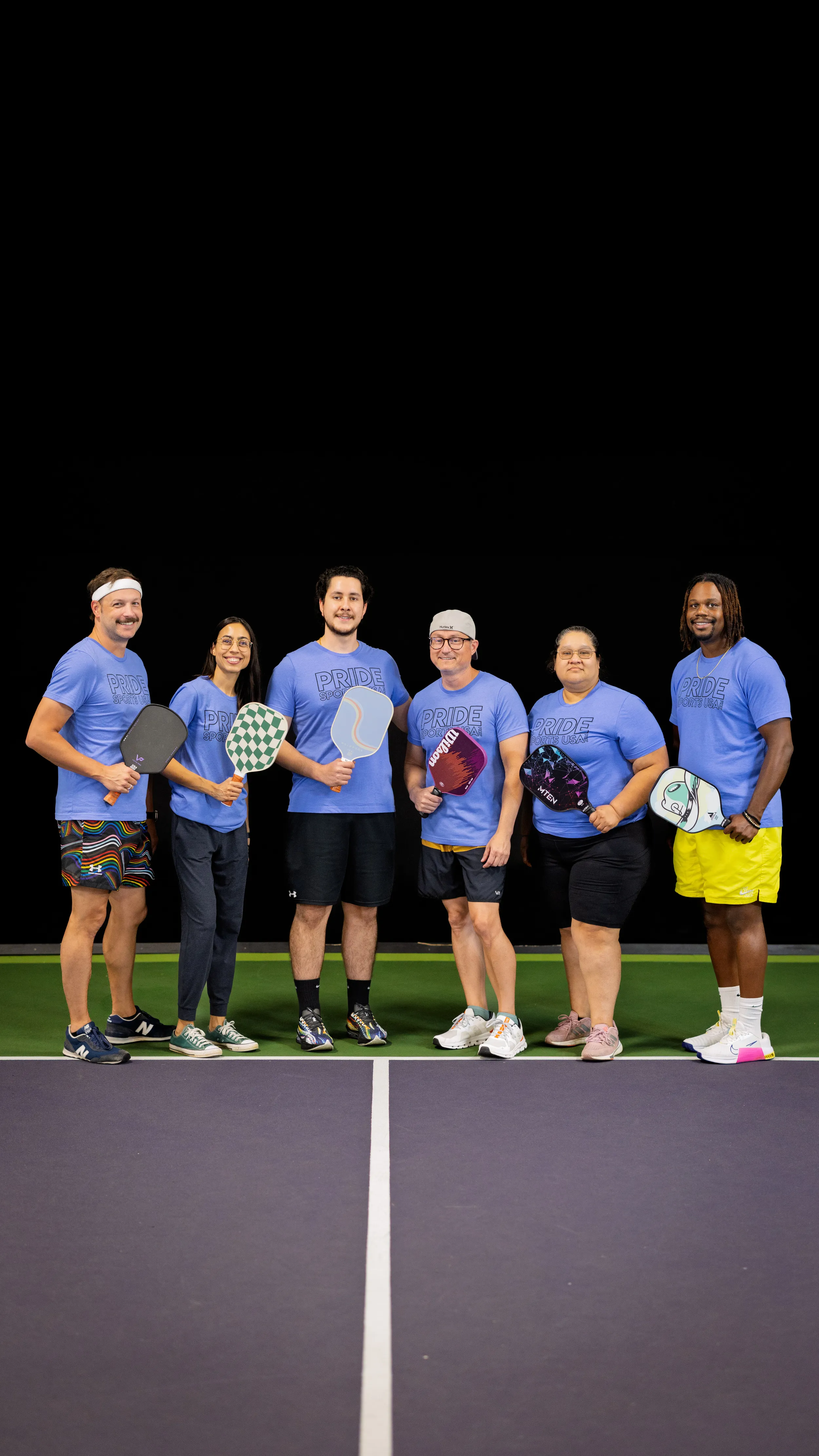 Six pickleball players pose at center court holding paddles with varied designs, wearing matching light-blue “PRIDE” shirts, sneakers, and athletic gear including a headband, rainbow-pattern shorts, and neon yellow shorts. The group stands on a lit pickleball court with clear boundary lines against a dark background.