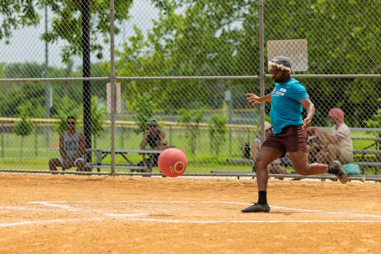 A player in a blue shirt, maroon shorts, and a headband is running on a kickball field, preparing to kick a red kickball as spectators watch from the benches behind a chain-link fence.